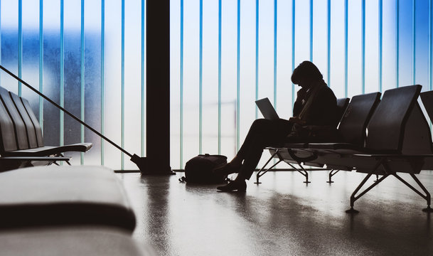 Business Woman Working In Empty Airport Terminal On Thin Laptop Computer Waiting For The Delayed Plane Preparing Working Documents Presentation Large Blue Glass Window