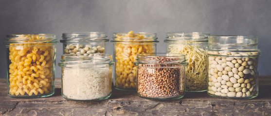 Various raw cereals, grains, beans and pasta for cooking healthy food in glass jars on a wooden table, on a gray background,  Clean food, vegan, balanced diet,zero waste,eco friendly,plastic free