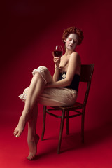 Medieval redhead young woman as a duchess in black corset and night clothes sitting on a chair on red background with a glass of wine. Concept of comparison of eras, modernity and renaissance.