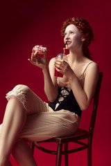Medieval redhead young woman as a duchess in black corset and night clothes sitting on a chair on red background with a drink and donut. Concept of comparison of eras, modernity and renaissance.