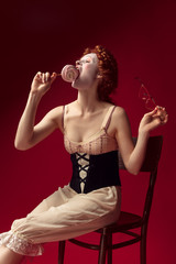 Medieval redhead young woman as a duchess in black corset, sunglasses and night clothes sitting on a chair on red background with a candy. Concept of comparison of eras, modernity and renaissance.
