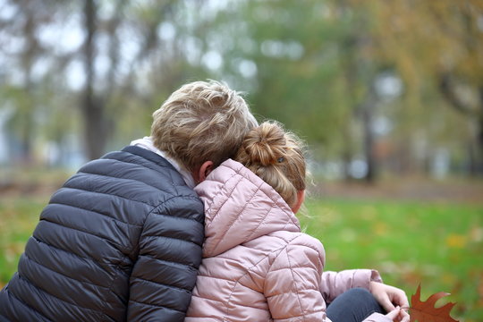 Mama z c&oacute;rką siedzą, objęte, przytulone, w parku, tyłem