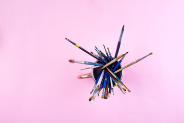 Paint brushes smeared in paint in a glass on a pink background.