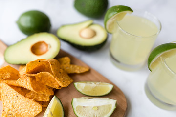 Fresh avocado, lime, drink and nacho chips lying on marble background. Recipe for Cinco de Mayo party