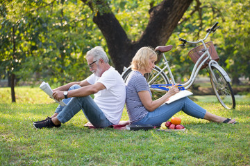 Happy senior couple relaxing sitting back to back in park reading newspaper and take write notes to Diary book  to together . old people in the summer park . Elderly resting .mature relationships .