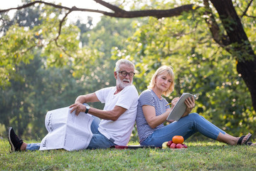 Fototapeta premium Happy senior couple relaxing sitting back to back in park reading newspaper and take write notes to Diary book to together . old people in the summer park . Elderly resting .mature relationships .