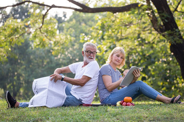 Happy senior couple relaxing sitting back to back in park reading newspaper and take write notes to Diary book  to together . old people in the summer park . Elderly resting .mature relationships .