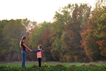Naklejka na ściany i meble Back view of young slim attractive mother and child girl standing in green meadow holding hands outdoors on forest trees blurred background.