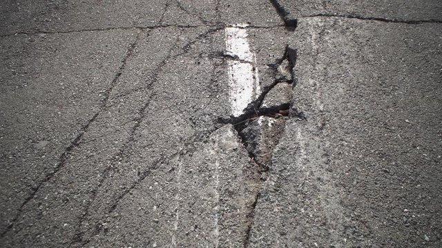 Camera moves along deep cracks in asphalt with white dashed marking line. View of fault in road, consequences of natural disaster, earthquake or man-made disaster.