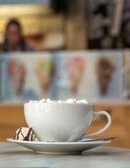 Mug of coffee with marshmallows on porcelain plate on blurred colorful interior bokeh background.