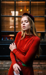 beautiful model with blond hair in an orange dress posing against the backdrop of large windows and looking away