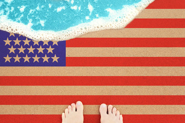 Two feet on the sunny sandy beach with flag USA. View from top on surf.