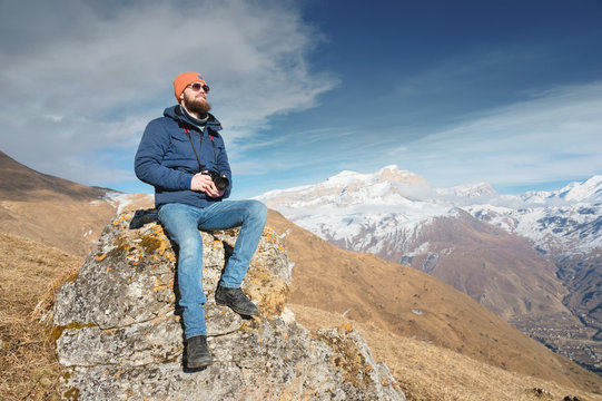 Portrait of a bearded traveler photographer in sunglasses and a cap sits on a rock with mirror camera in his hands against the backdrop of mountains
