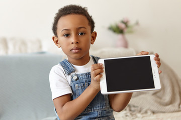 Happy childhood, ethnicity, rest and relaxation. Happy adorable eight year old African boy lying on stomach on carpet at home, keeping hands on his cheeks, smiling joyfully as if watching cartoons