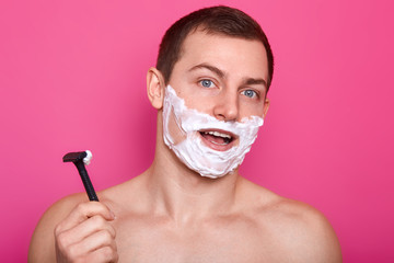 Studio shot of attractive guy posing with razor in bathroom, standing with cream on face and opened mouth, has surprised facial expression, isolated over pink background. Skin and body care concept.