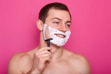 Happy handsome man shaving his face over rose background, prepares for dating, looking at mirror with charming smile, posing with foam on his cheeks, keeps razor in hand. Everyday procvedures concept.