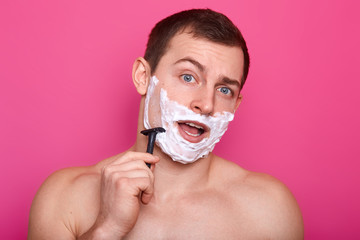 Image of young man posing with open mouth, shaving in bathroom and singing, keeps razor in hand, stands with naked shoulders, doing everyday procedures, isolated on rose background. Skin care concept.