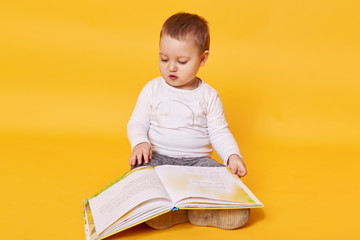 Toddler girl pretends to read book while sitting on floor, viewing pictures and turning pages, little girl looks concentrated, wearing casual white shirt, posing isolated over yellow background.