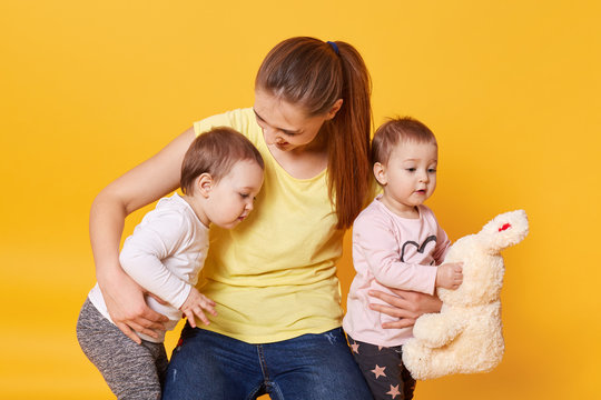 Image of mother with kids, playing with mommy while posing in photo studio, girl holds plushy rabbit, toddlers dressed in casual clothes, celebrating Mother's Day, isolated over yellow background.