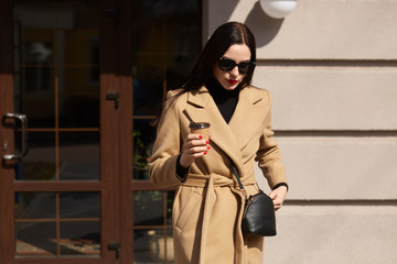 Outdoor shot of beautiful elegant woman wearing beige coat and black suglasses, goes along street...