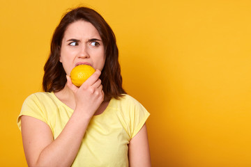 Natural vitamins and healthy food concept. Indoor shot of young brunette woman bits lemon with discontented facial expession, looking angrily aside, copy space for your promotion text or advertisment.