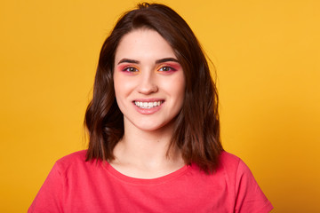 Close up portrait of beautiful caucasian young woman with pink and orange glamour make up and toothy smile, model has happy facial expression, dressed casually, posing isolated on yellow background.