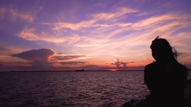 Woman sitting looking at sunset sky at sea beach . Woman silhouette backside view watches evening ocean waves enjoying wind at her face thinking dreaming