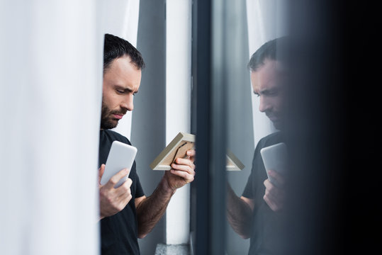 selective focus of depressed handsome man holding smartphone and photo frame while standing by window