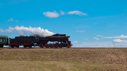 Fototapeta na wymiar The locomotive rides on the way to a high mound. From the pipe of the locomotive is white steam. There are white clouds in the blue sky.