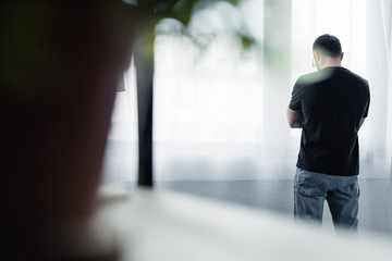 selective focus of depressed man in black t-shirt standing by window at home