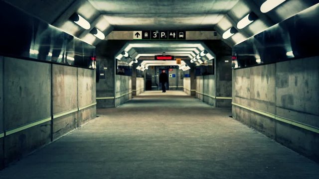 A dark tunnel with a man walking through alone at night.One man, alone, walking through an empty concrete subterranean pedestrian tunnel  lines with lights and reflective metal. It is under a GO train