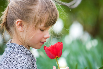 Profile of cute pretty smiling child girl with gray eyes and long hair smelling bright red tulip flower on blurred sunny summer green bokeh background.