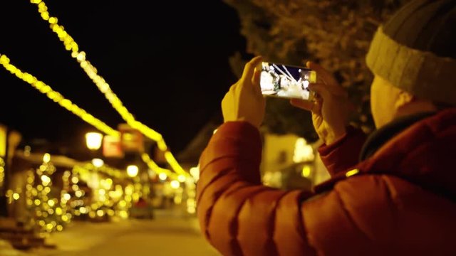 Unknown young man in red jacket makes photos with smartphone in the evening on Christmas illumination background. Shot on Red camera