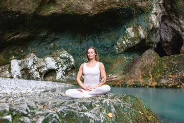 Inspired young beautiful female and yoga coach meditating sitting in a white suit in the lotus position with eyes closed in the wild nature against the background of the river and green hills