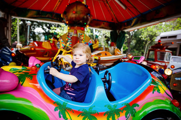 Obraz na płótnie Canvas Adorable little toddler girl riding on funny car on roundabout carousel in amusement park. Happy healthy baby child having fun outdoors on sunny day. Family weekend or vacations