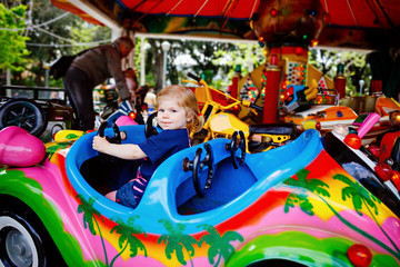 Adorable little toddler girl riding on funny car on roundabout carousel in amusement park. Happy...
