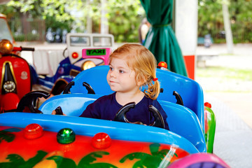 Adorable little toddler girl riding on funny car on roundabout carousel in amusement park. Happy healthy baby child having fun outdoors on sunny day. Family weekend or vacations