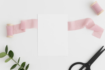 Top view of a white card mockup with silk ribbons, scissors and green branch on a white table.