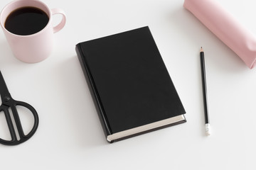 Top view of a black book mockup with workspace accessories and a cup of coffee on a white table.