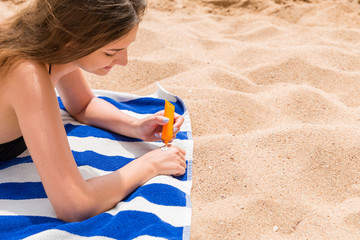 Attractive woman is lying on a striped towel at the beach and squeezing sunblock from a tube on her hand