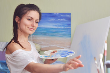 A beautiful woman artist paints a picture of blue acrylic paints on a large white canvas at her home against the background of her painting of the sea.