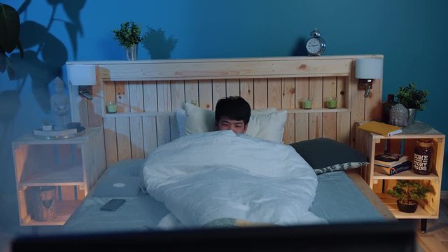 Tired young asian man fells asleep in comfortable bed. Good night, dreams, relaxation. Beautiful wooden bed, cozy lights and accessories. Having a rest. Comfort concept