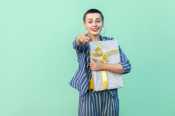 Winner are you! Portrait of happy beautiful with short hair young woman in striped suit standing, hugging present box and pointing finger to camera. Indoor, isolated, studio shot, green background