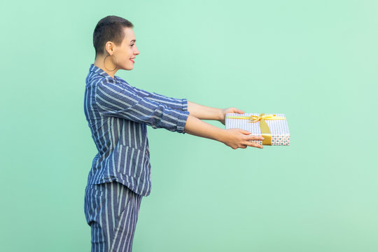 Congratulation! Side view portrait of beautiful with short hair young woman in striped suit standing, giving you present box, with toothy smile. Indoor, isolated, studio shot, green background - Powered by Adobe