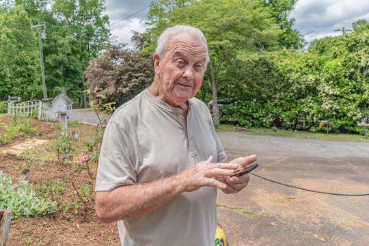 A Senior Man Enjoys Checking Is Email And Text Messages And Social Media On His Phone While In His Garden On A Nice Day.