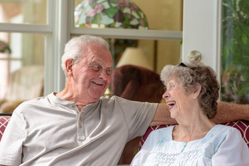 A beautiful senior couple in their seventies laughs while enjoying time together on their porch on a sunny day.