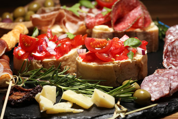 Traditional italian antipasto bruschetta appetizer with cherry tomatoes, cream cheese, basil leaves and balsamic vinegar on cutting board with prosciutto, salami, cheese