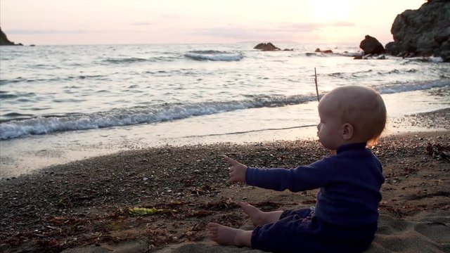Adorable caucasian 1,5 years old boy is sitting on sand, looking at splashing sea waves on the beach and playing with sticks. Ritz bay, Nakhodka, Russia. September evening