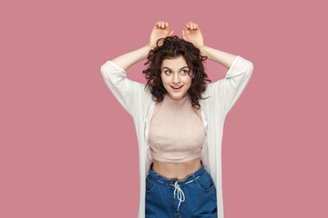 Portrait of funny happy cute brunette young woman with curly hairstyle in casual style standing with hands on head in bunny gesture and looking away. indoor studio shot isolated on pink background.