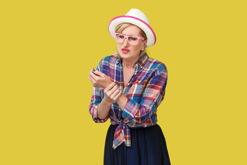 Pain on hand or wrist. Portrait of sick stylish mature woman in casual style with hat and eyeglasses standing and holding her painful wrist hand. indoor studio shot isolated on yellow background.